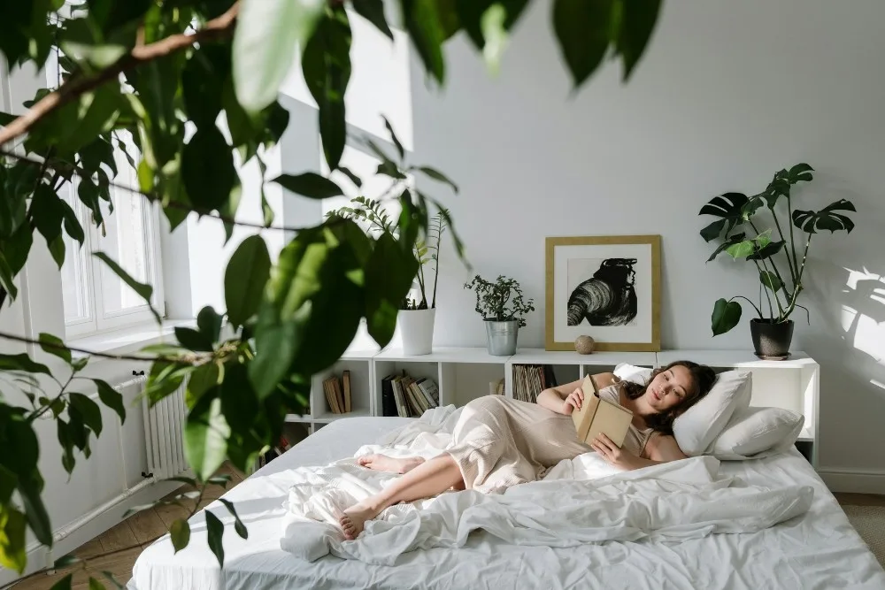 Cô gái tiết kiệm chi tiêu nằm đọc sách trên giường trong phòng ngủ bố trí nhiều cây xanh