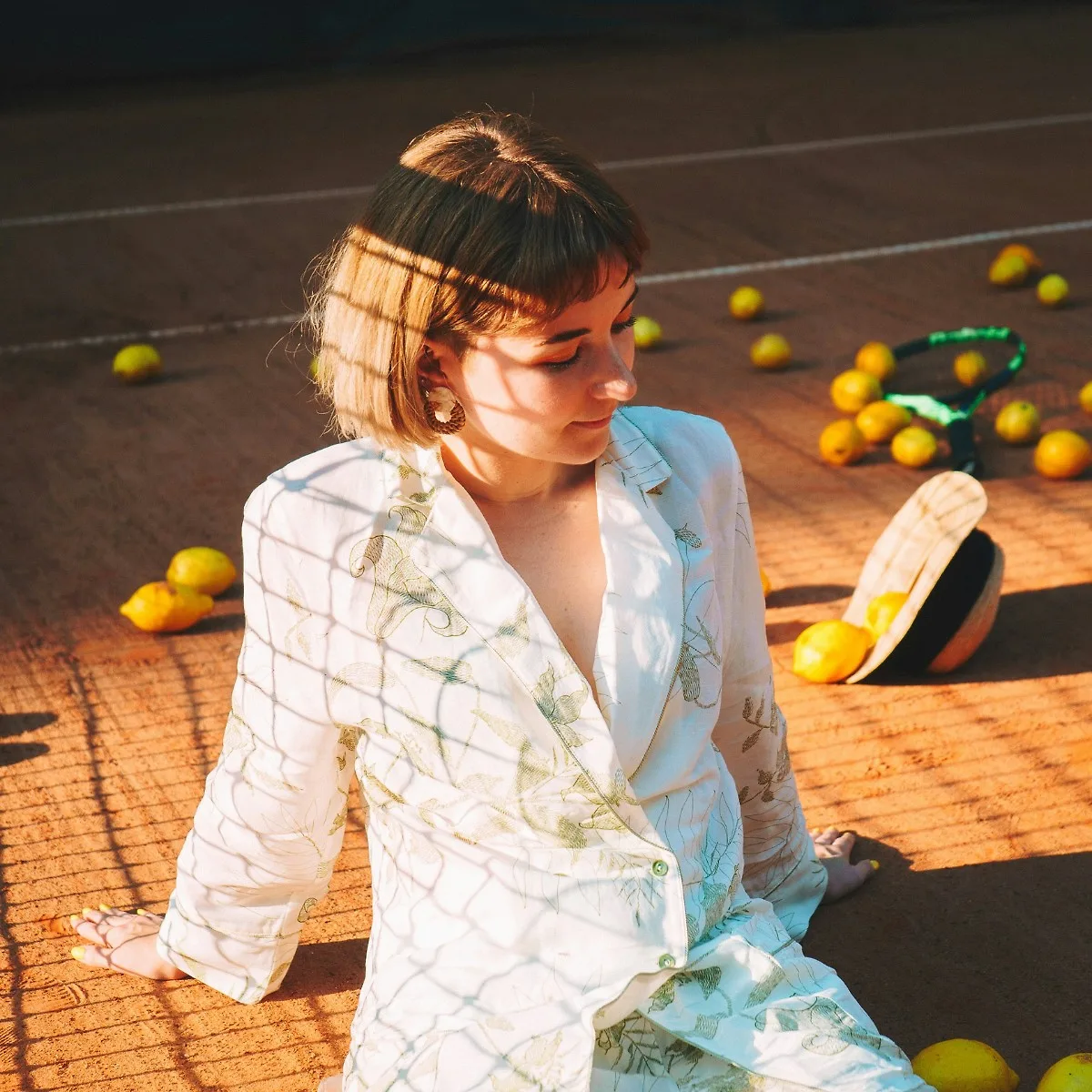 Cô gái tóc ngắn mặc sơ mi trắng ngồi trên sân tennis với những quả chanh vàng