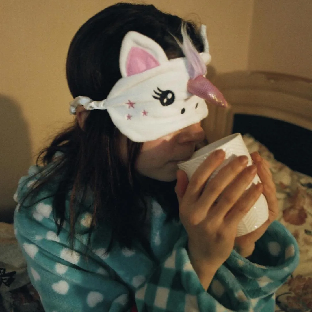 Cô gái mặc áo ngủ dễ thương, đeo bịt mắt ngủ hình unicorn, đang cầm ly nước nóng.