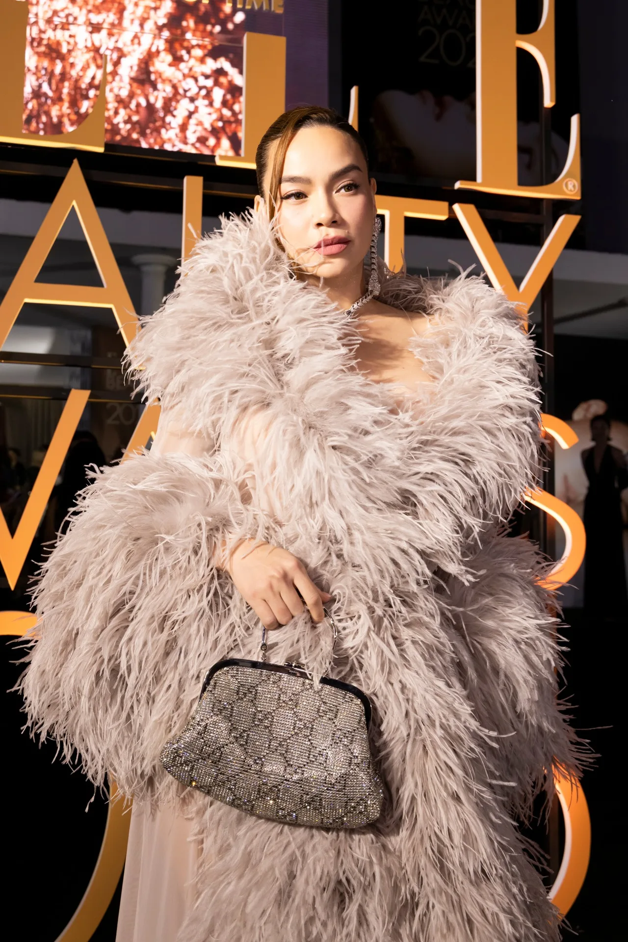 Xuất hiện  trên thảm đỏ sự kiện ELLE Beauty Awards 2025, Ca sĩ Hồ Ngọc Hà lựa chọn thiết kế lông vũ đến từ BST La Famiglia của Gucci, đi cùng túi xách Crystal Medium Evening Top Handle Bag kết hợp hoa tai và vòng cổ Dionyus. Hồ Ngọc Hà hoàn thiện với kiểu tóc slick-back rẽ ngôi lệch, giúp gương mặt và phần cổ cao của thiết kế trở nên sắc sảo hơn. Lớp makeup tông nude-glowy tập trung vào làn da căng bóng, eyeliner mắt mèo và đôi môi beige hồng tạo cảm giác sang trọng, hiện đại nhưng vẫn đầy quyền lực.