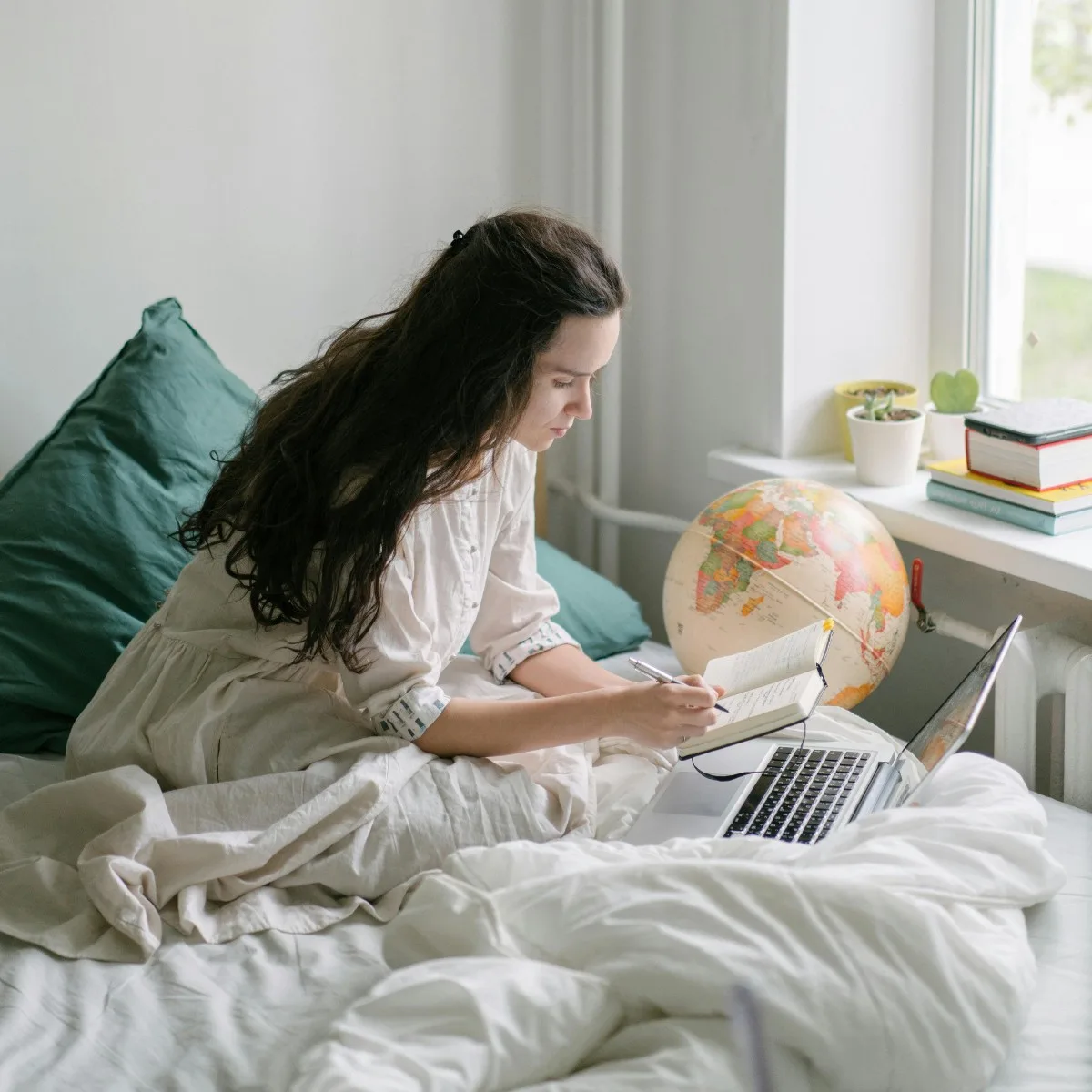 Cô gái tóc dài ngồi ghi chép bên laptop và quả địa cầu ở trên giường ngủ