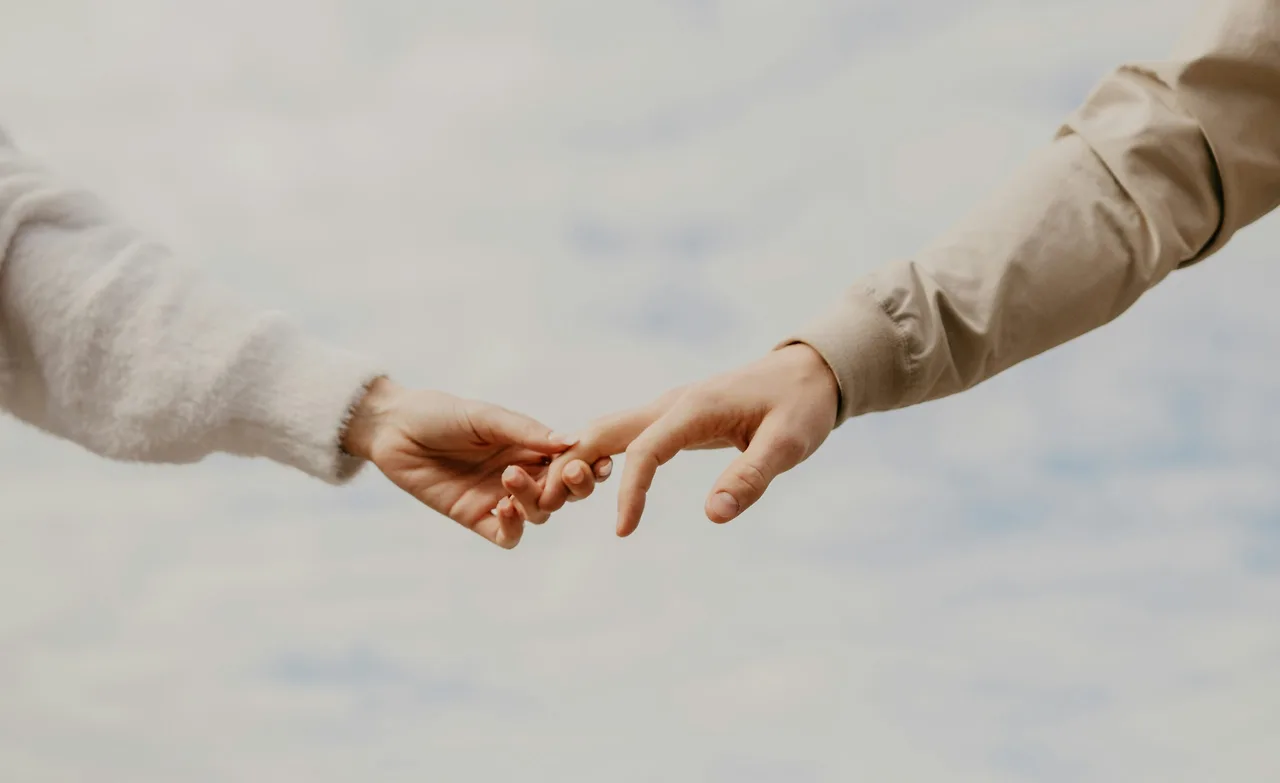 Góc máy cận cảnh tập trung vào đôi bàn tay của hai người đang khẽ chạm và tìm đến nhau trên nền trời mây nhạt. Hành động đan tay này là một dấu hiệu xoa dịu và truyền đi sự đồng cảm mạnh mẽ.