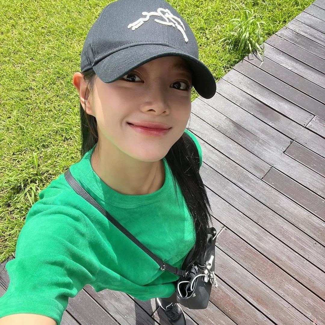 Cô gái đội mũ lưỡi trai đen, mặc áo thun xanh lá và đeo túi chéo, selfie ngoài trời với nụ cười rạng rỡ dưới ánh nắng - ngăn ngừa lão hóa