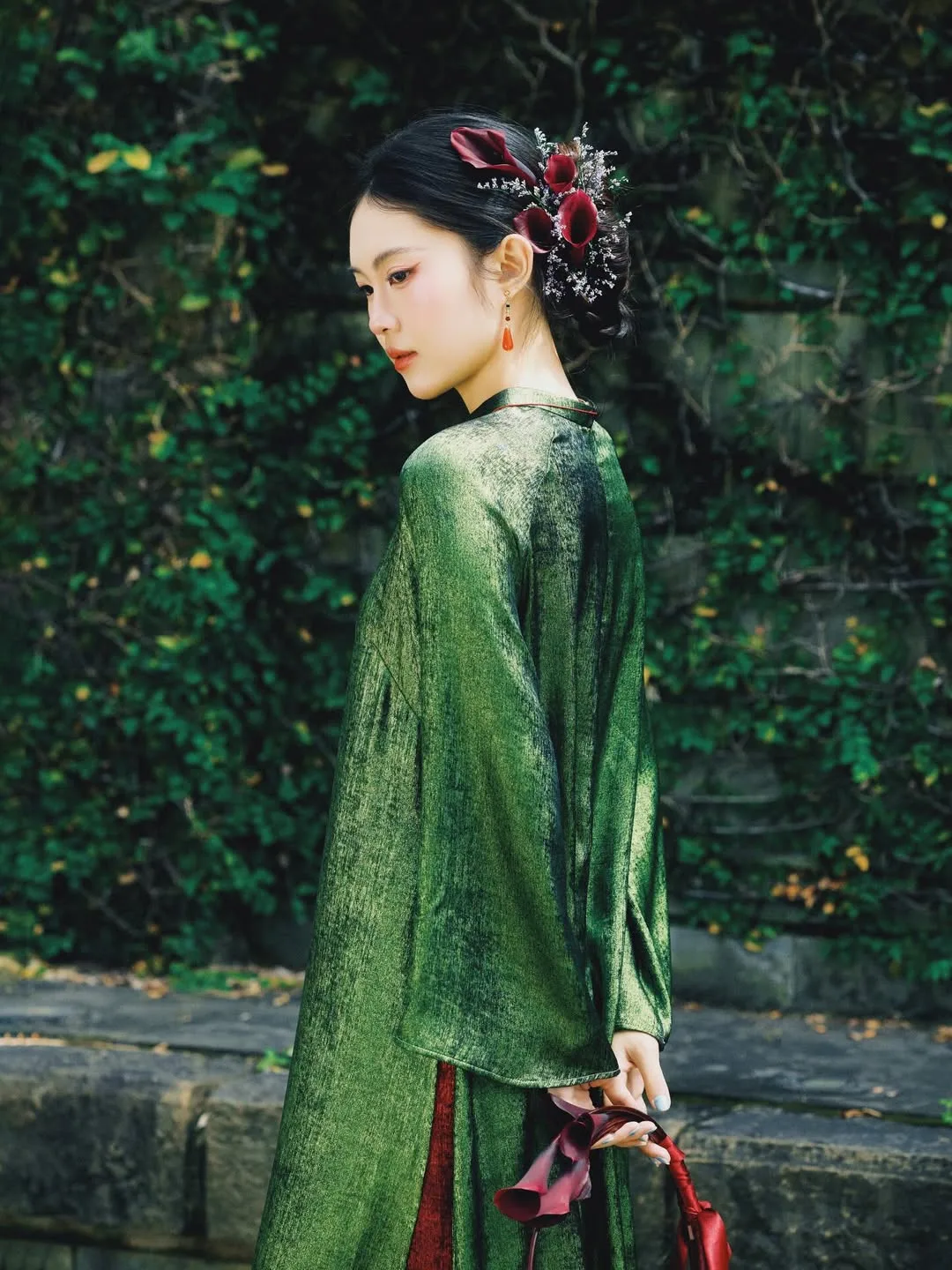 Một chiếc kẹp hoa trà đỏ rực cài lệch trên mái tóc búi thấp, tạo điểm nhấn tương phản trên nền áo dài lụa màu xanh ngọc.