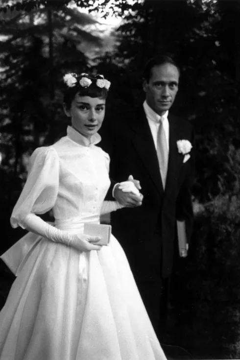 Năm 1954, khi kết hôn lần đầu với Mel Ferrer, minh tinh màn bạc Audrey Hepburn chọn chiếc váy trắng, độ dài đến mắt cá và “kín cổng cao tường”. Điểm nhấn của bộ váy là hai tay được thiết kế bồng và những bông hoa điểm xuyết trên mái tóc theo phong cách vũ công ballet.