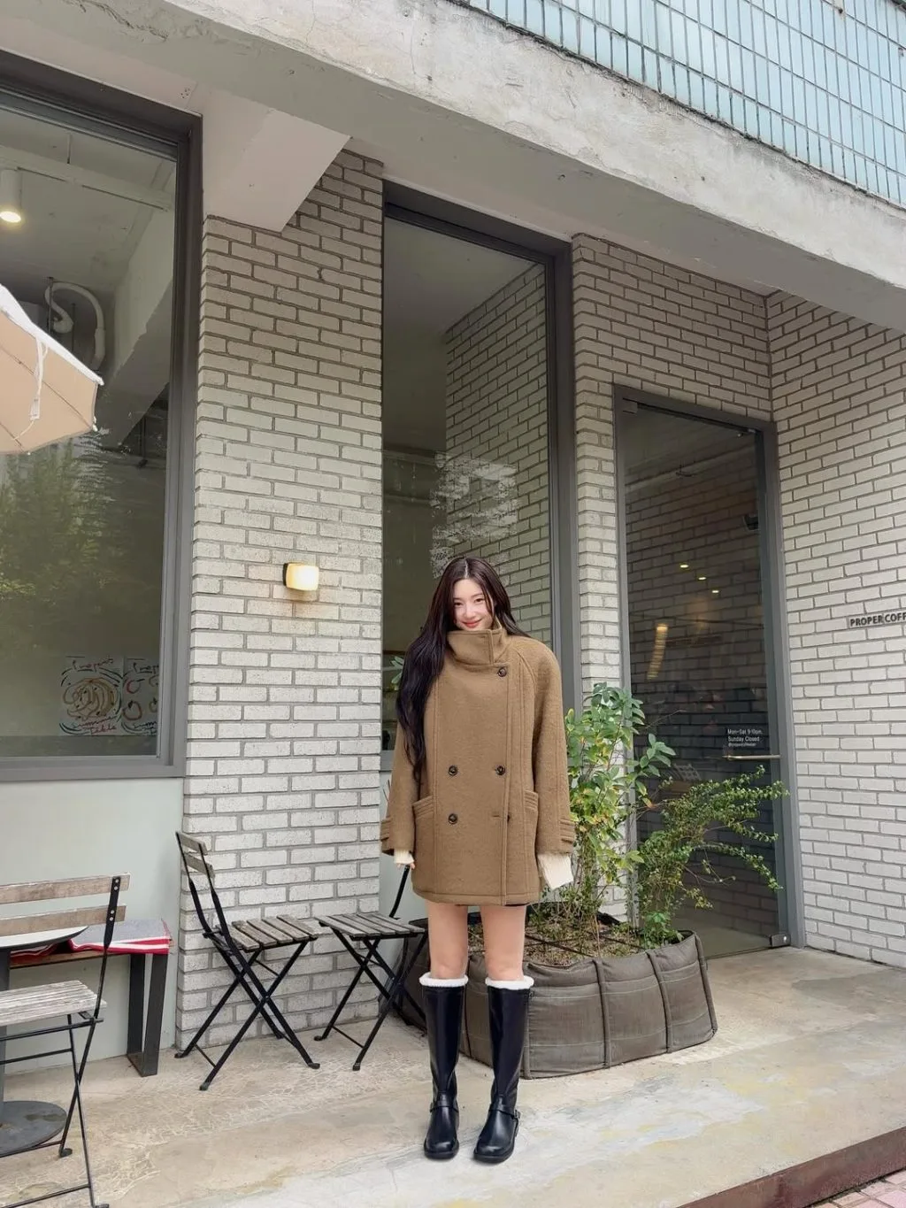 Cô gái khoác áo măng tô camel bước đi giữa phố, đôi giày boots ánh đen tạo điểm nhấn cổ điển.