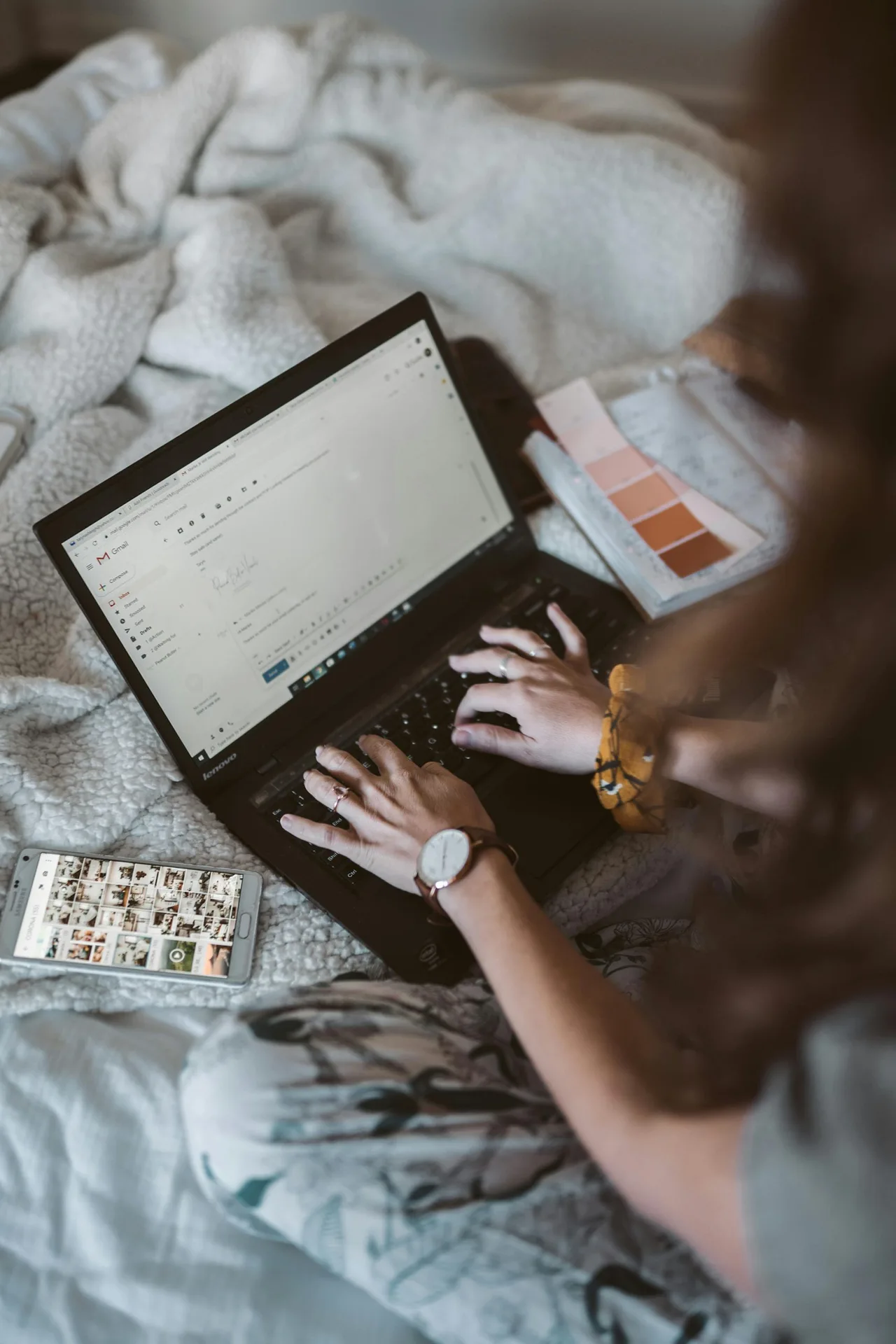 cô gái ngồi trên giường check mail trên máy tính bên cạnh điện thoại và bảng màu