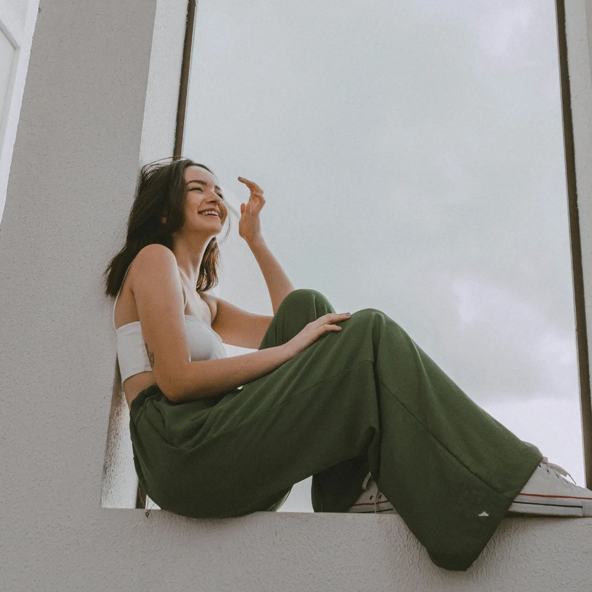 Cô gái tóc đen mặc quần dài màu xanh lá ngồi trên ô cửa tươi cười có dấu hiệu tích cực
