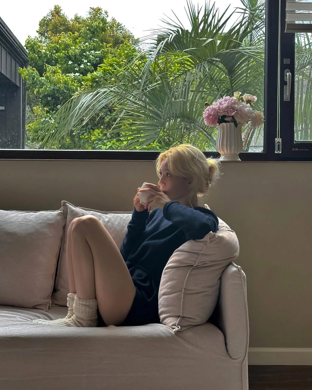 Somi mặc áo màu xanh navy tóc blonde ngồi trên sofa uống nước vào buổi sáng, thanh lọc