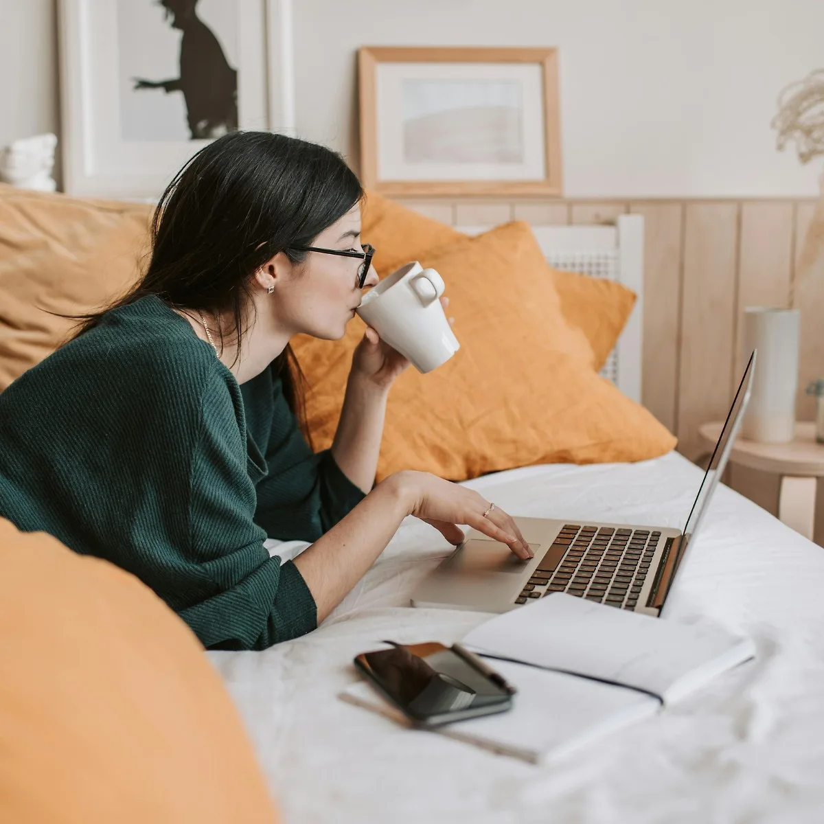 Cô gái tóc đen đeo mắt kính uống cà phê làm việc bên laptop trên giường ngủ