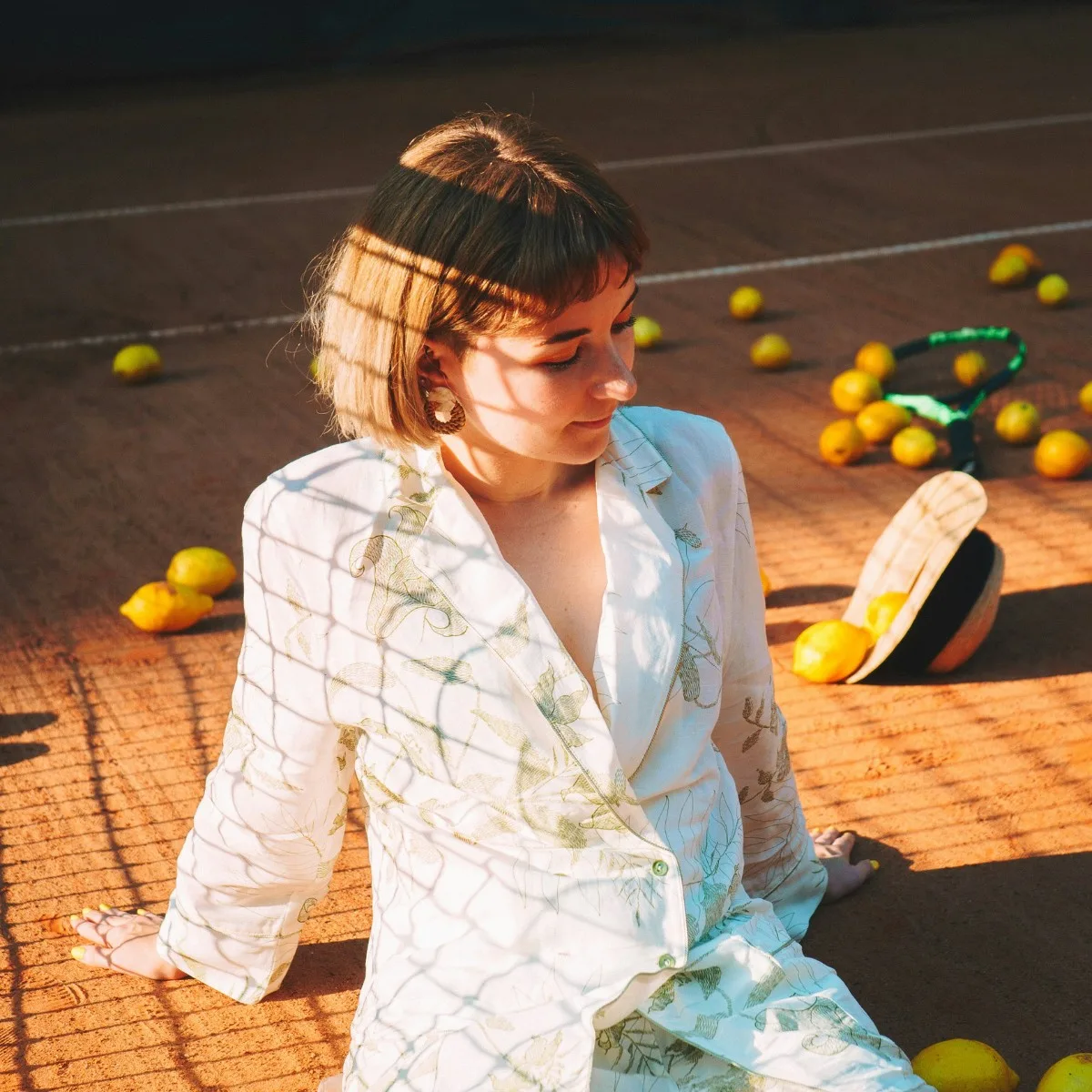 Cô gái tóc ngắn mặc sơ mi trắng ngồi trên sân tennis với những quả chanh vàng
