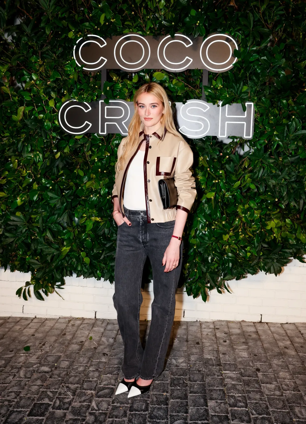 Kathryn Newton đầy trẻ trung khi diện áo khoác da màu beige viền nâu nổi bật, kết hợp cùng quần jeans xám khói và túi cầm tay đen sang trọng, tạo nên một tổng thể vừa năng động vừa thời thượng.